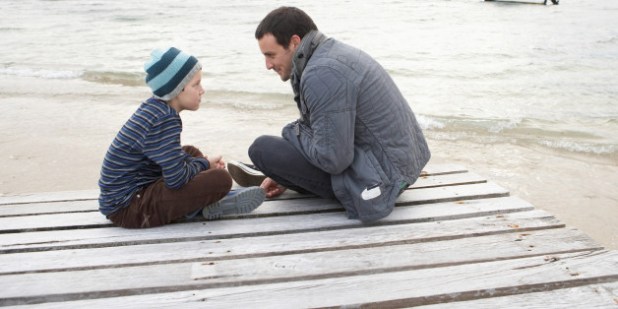 Father and son (10-11) sitting at end of dock at edge of lake, talking, side view